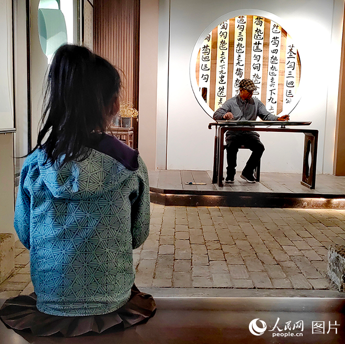 在西河古镇弋農古琴斫制工艺馆内，一位小朋友正在全神贯注地聆听弋農古琴非物质文化遗产代表性传承人翟光宝弹奏的古琴曲。翟光宝是弋農古琴斫制工艺的第四代传承人。弋農古琴又称“百纳琴”，是以古木片合漆胶合而成的琴，始创于唐代。