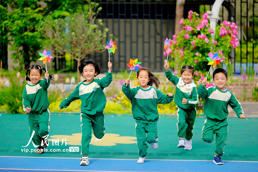 2025年5月8日，在江苏省南通市，崇川区沁园幼儿园的小朋友手持小风车在操场上微笑着奔跑。徐培钦摄（人民图片网）