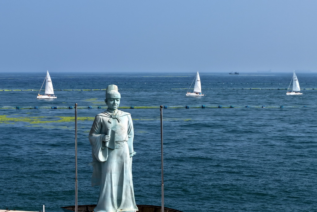 青岛银海大世界港池外,参加海上巡游的帆船从郑和雕像旁驶过(7月10日摄,无人机照片)。